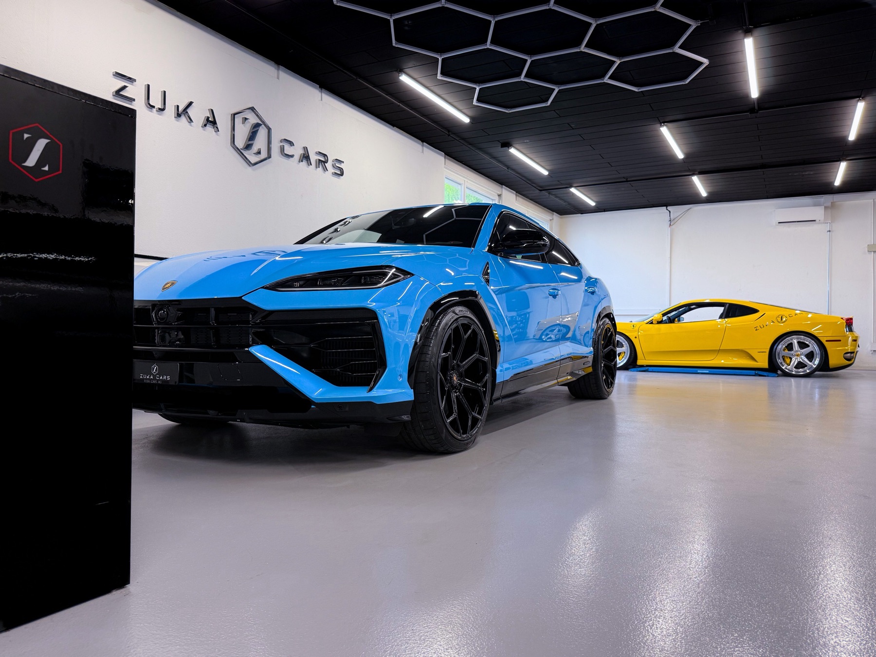 Blauer Lamborghini Urus und gelber Ferrari in der Werkstatt von Zuka Cars, an der schwarzen Decke die Waben-Leuchten